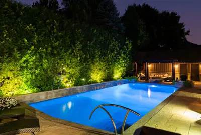 Conscape Lighting + Audio Rectangular swimming pool illuminated at night with elegant Toronto lighting, surrounded by trees and lounge chairs, and featuring a covered seating area in the background.
