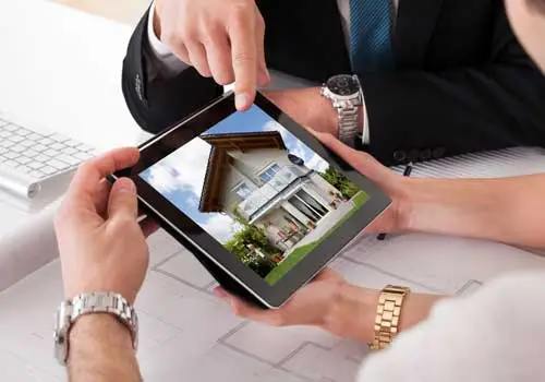 Conscape Lighting + Audio Two people viewing a photo of a house with Conscape Lighting + Audio’s outdoor lighting on a tablet, alongside architectural plans and a keyboard, planning their next Toronto lighting project.