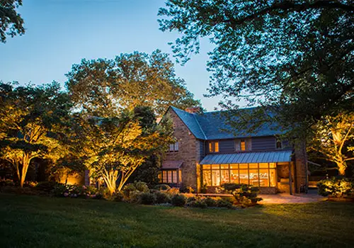 Conscape Lighting + Audio Two-story house with large windows is lit up at dusk by Conscape Lighting + Audio, surrounded by trees and a landscaped garden with a well-kept lawn.