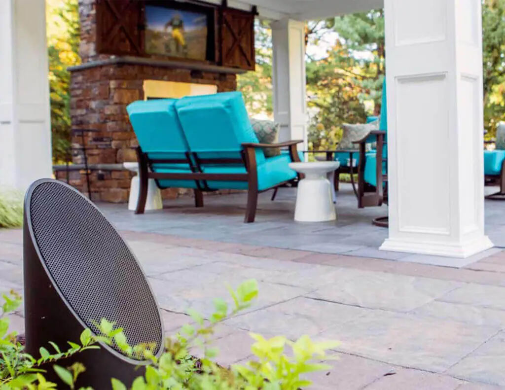Conscape Lighting + Audio Outdoor seating area with blue cushioned chairs, a TV above a stone fireplace, and a black outdoor speaker in the foreground surrounded by plants—experience immersive outdoor audio with Conscape Lighting + Audio solutions.