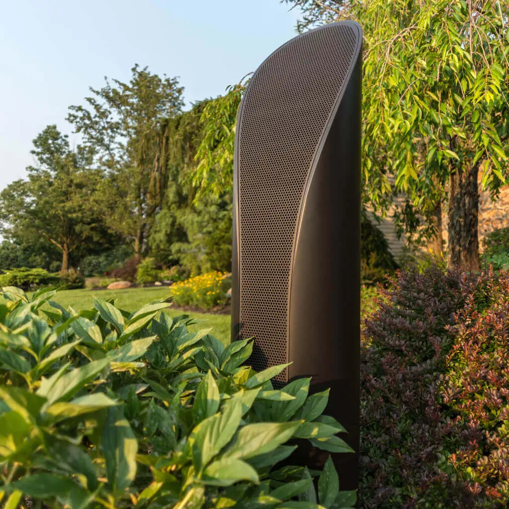 Conscape Lighting + Audio A tall, modern outdoor speaker stands among green plants and bushes in a landscaped garden, complementing the outdoor lighting for a perfect blend of style and outdoor audio.