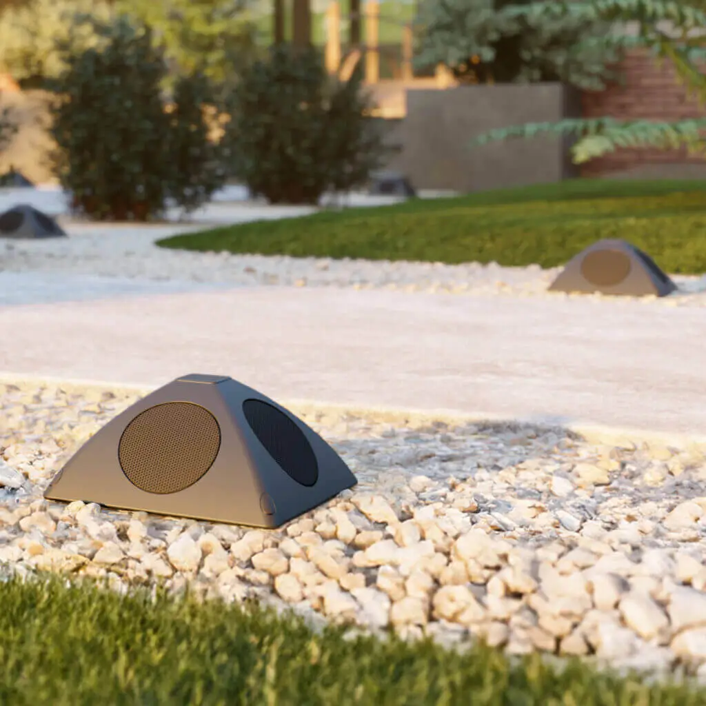 Conscape Lighting + Audio Black outdoor speakers from Conscape Lighting + Audio are installed on white gravel along a garden pathway, with green grass and plants in the background, enhancing your outdoor lighting and audio experience.