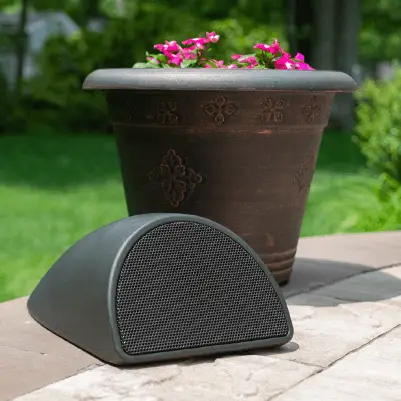 Conscape Lighting + Audio A black Conscape Lighting + Audio outdoor speaker sits on a stone surface in front of a large brown flowerpot with pink flowers, with greenery and outdoor lighting softly illuminating the background.