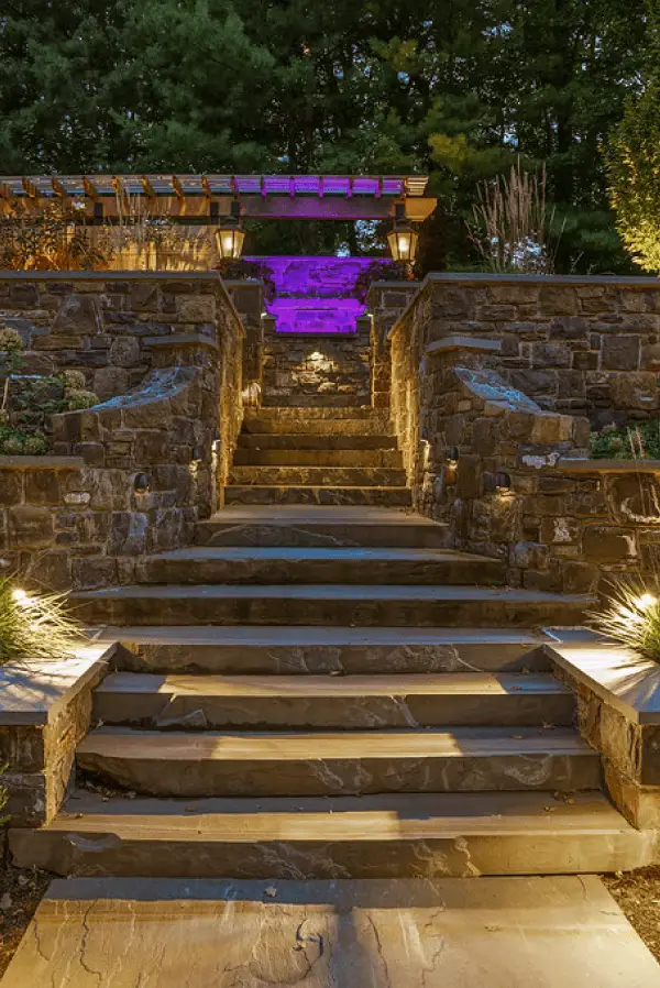 Conscape Lighting + Audio Stone steps lead to a terrace with stone walls, outdoor lighting from Conscape Lighting + Audio, and a pergola with purple illumination in the background, all surrounded by lush greenery.