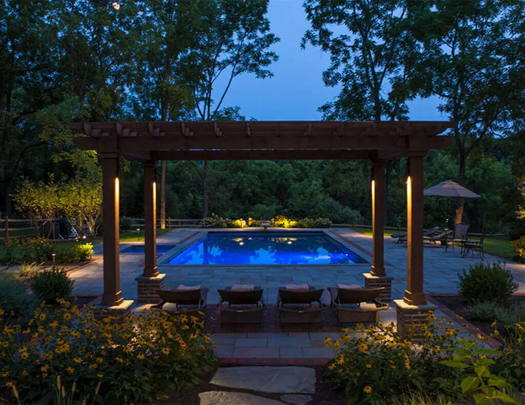 Conscape Lighting + Audio A backyard pool area at dusk, featuring a wooden pergola with Conscape Lighting + Audio, cozy lounge chairs, stone patio, and surrounding gardens with trees softly illuminated in the background.
