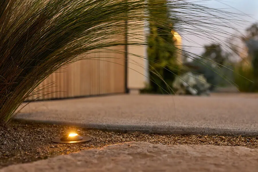 Conscape Lighting + Audio A small ground light from GTA Lighting is illuminated next to ornamental grass and a stone, beside a concrete walkway outside a modern building.
