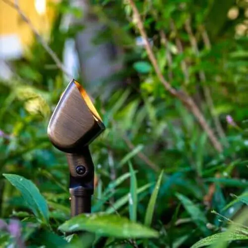 Conscape Lighting + Audio Outdoor landscape spotlight by Conscape Lighting + Audio is partially hidden among green plants, facing slightly upward, with a blurred background—perfect for integrating outdoor audio seamlessly into your space.