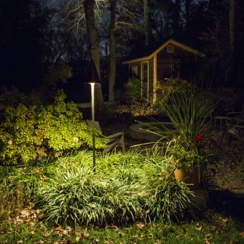 Conscape Lighting + Audio A garden at night glows under Conscape Lighting + Audio's outdoor lighting, highlighting lush plants and bushes in the foreground and a small shed in the background.